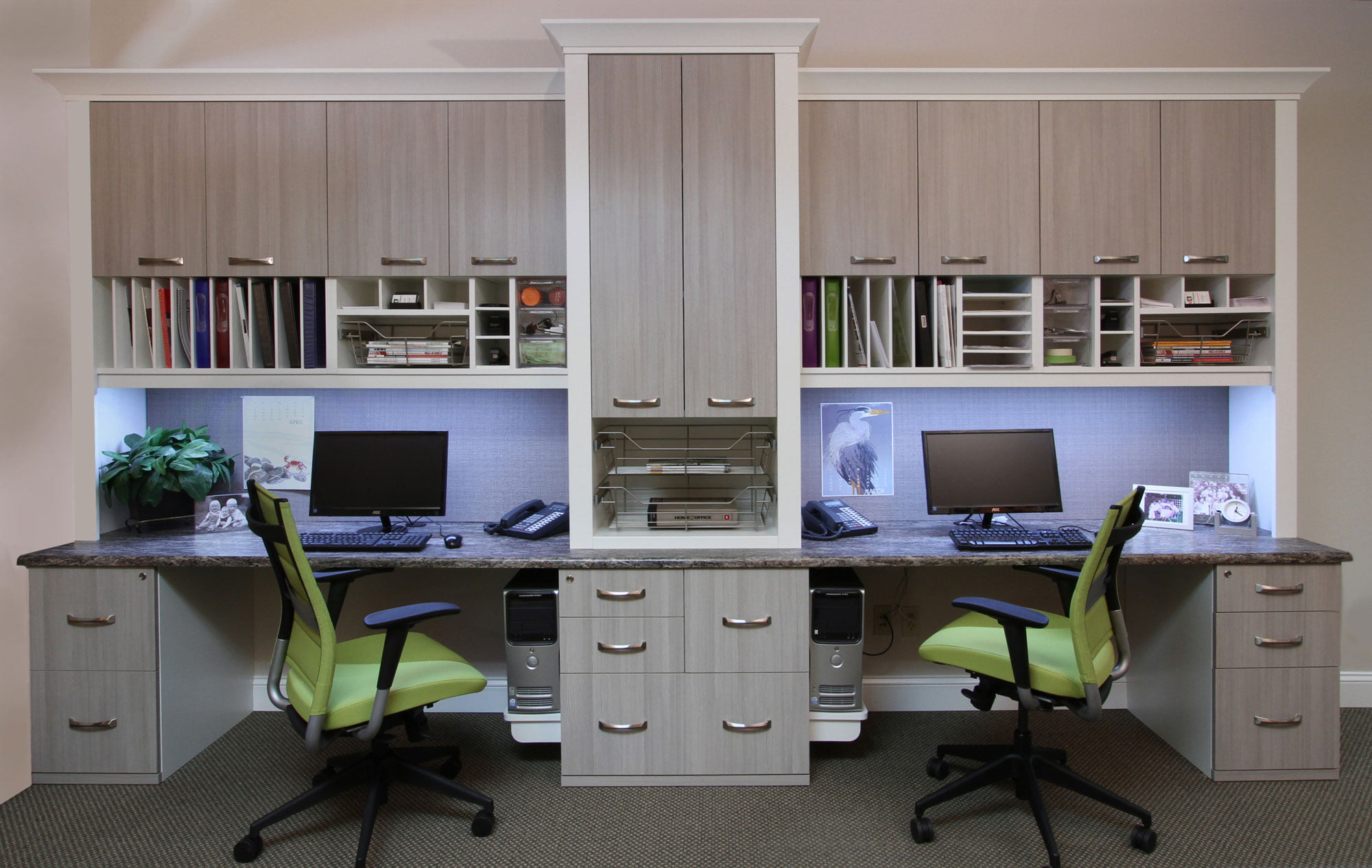 Organized home office with light wood cabinets, desks, filing drawers, computers, and green chairs.