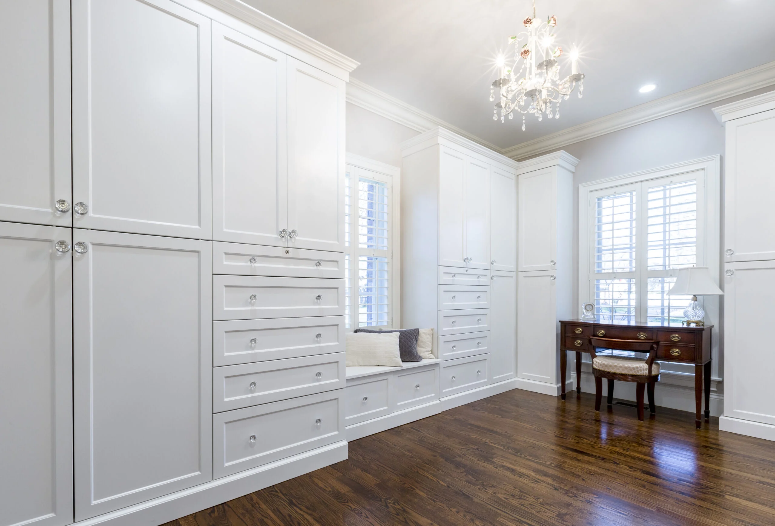 A bright, luxurious room with custom white built-in cabinetry, a window seat, dark wood floors, and a crystal chandelier.