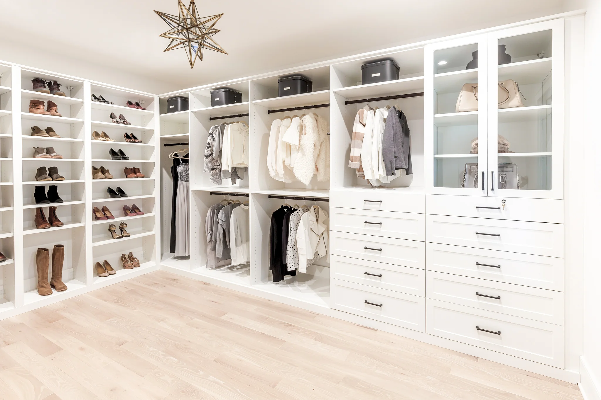 Bright, modern white walk-in closet with shelves for shoes, hanging clothes, drawers, and glass-door cabinets.