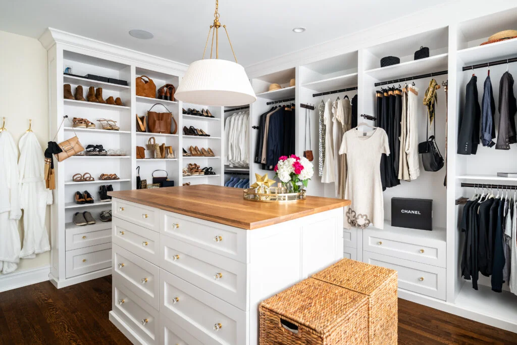 Spacious white walk-in closet with shoe shelves, hanging clothes, center island with wood top, and gold accents.