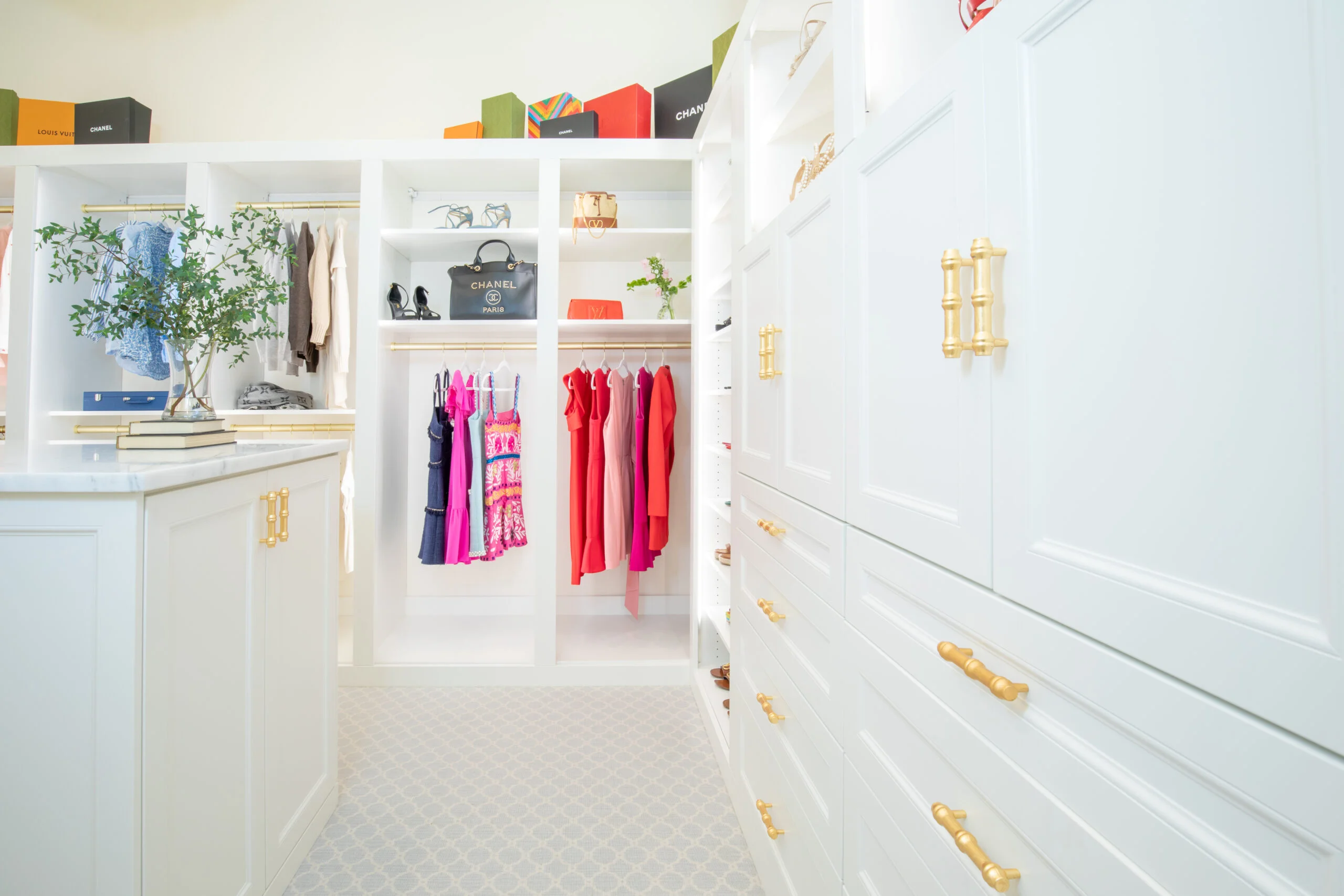 Spacious white closet with golden hardware, clothes on racks, shelves with designer bags and shoes, and a patterned rug.