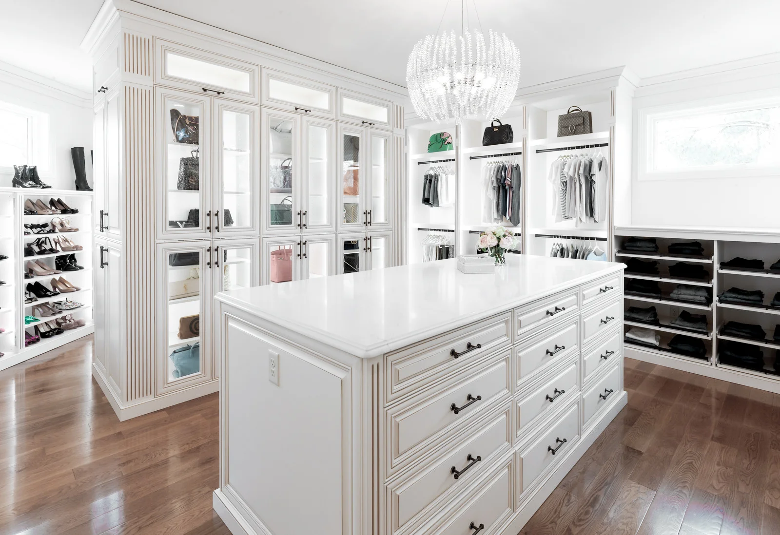 White custom closet with center island, glass-front cabinets for bags, shelves for shoes, and hanging clothes.