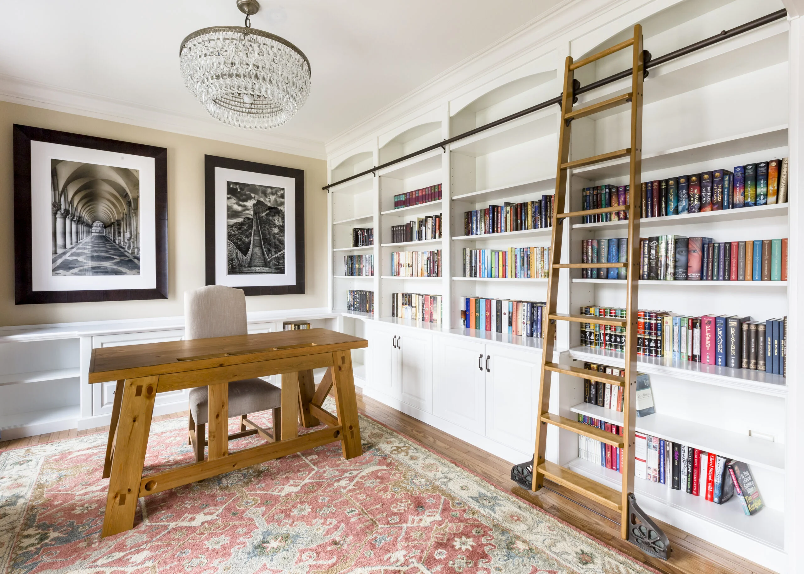 A home office features a wooden desk on a patterned rug, a cushioned chair, and white built-in bookshelves with a rolling ladder.