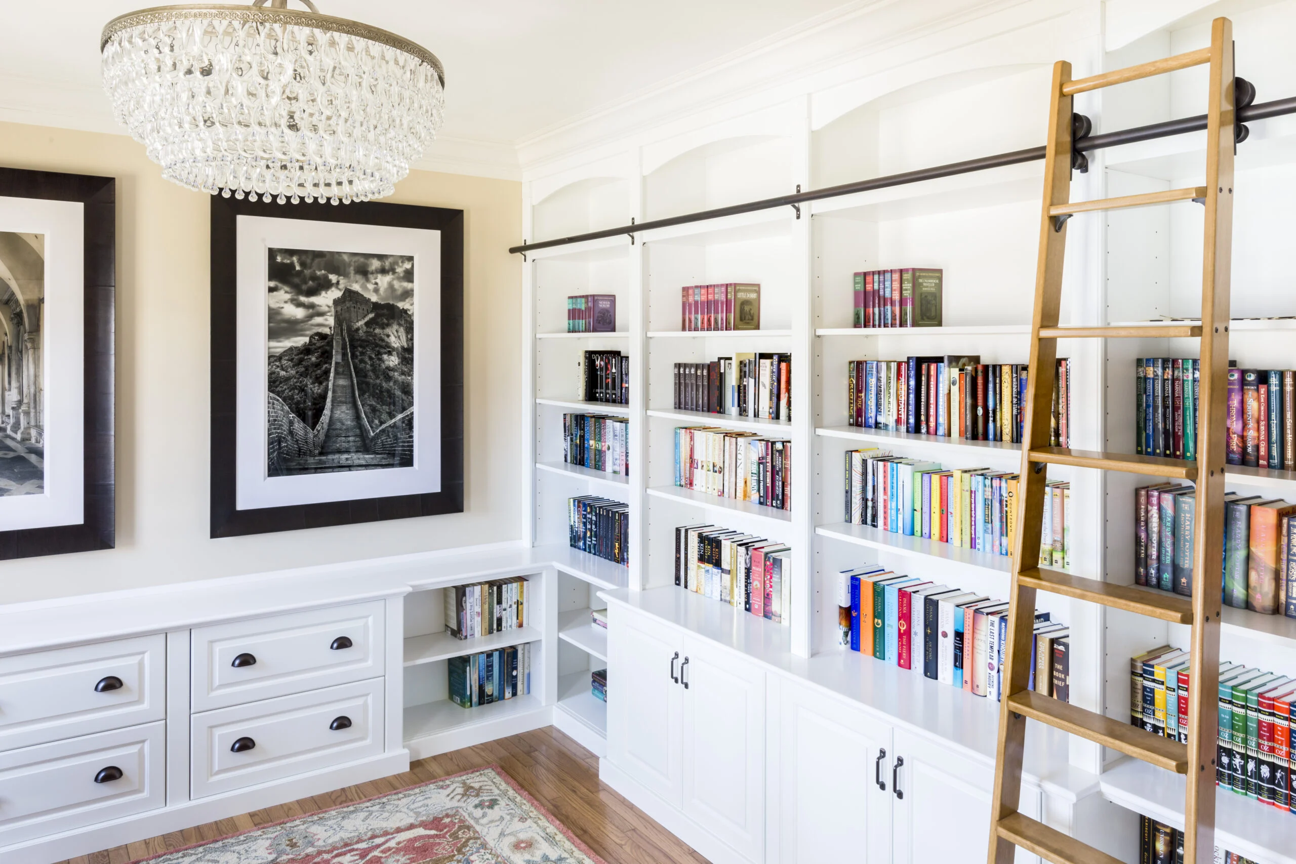 Elegant home library with white built-in bookshelves, various books, rolling ladder, and framed artwork.