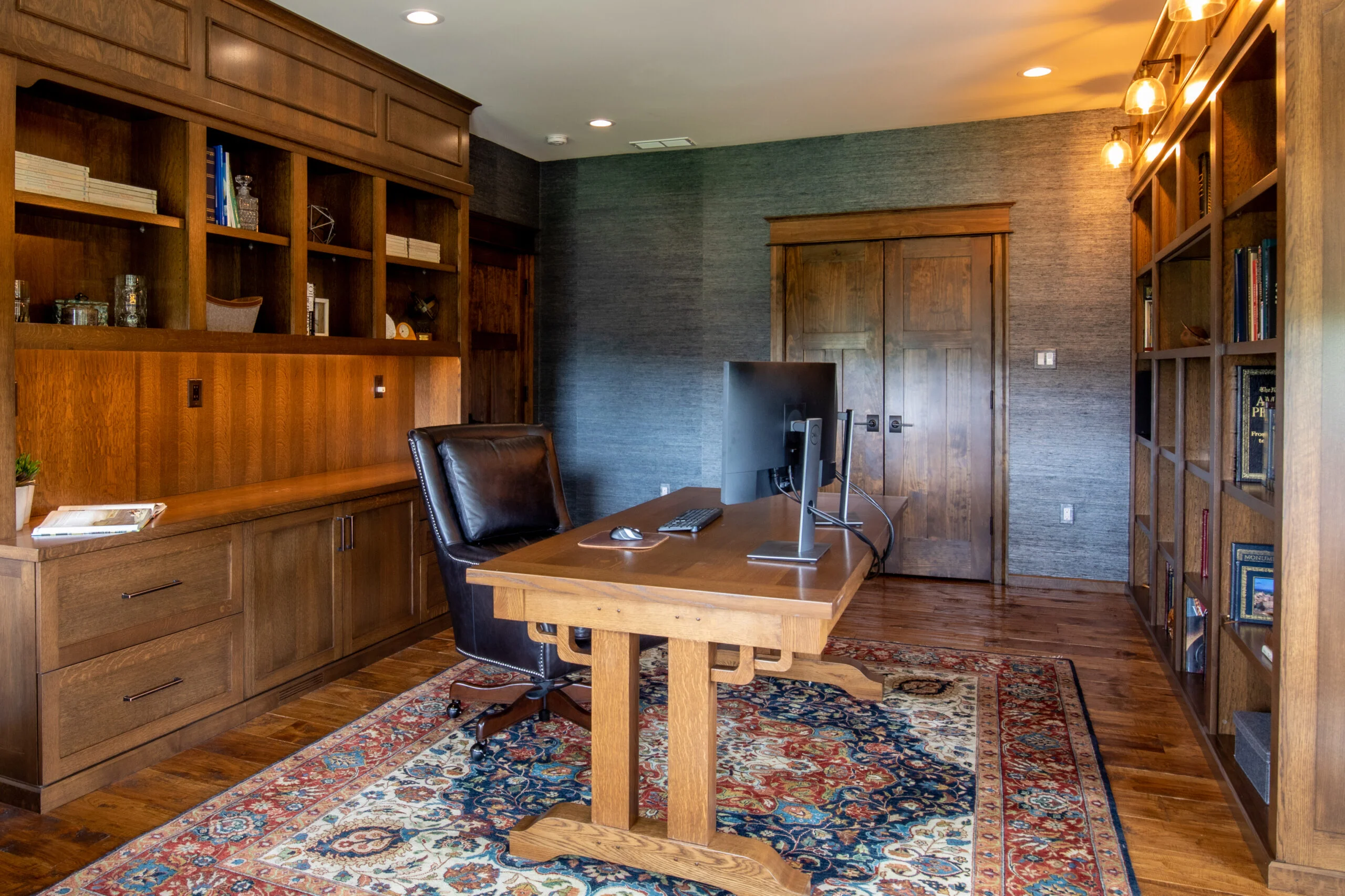A home office with dark wood built-in bookshelves, an executive desk, a leather chair, and a patterned rug.