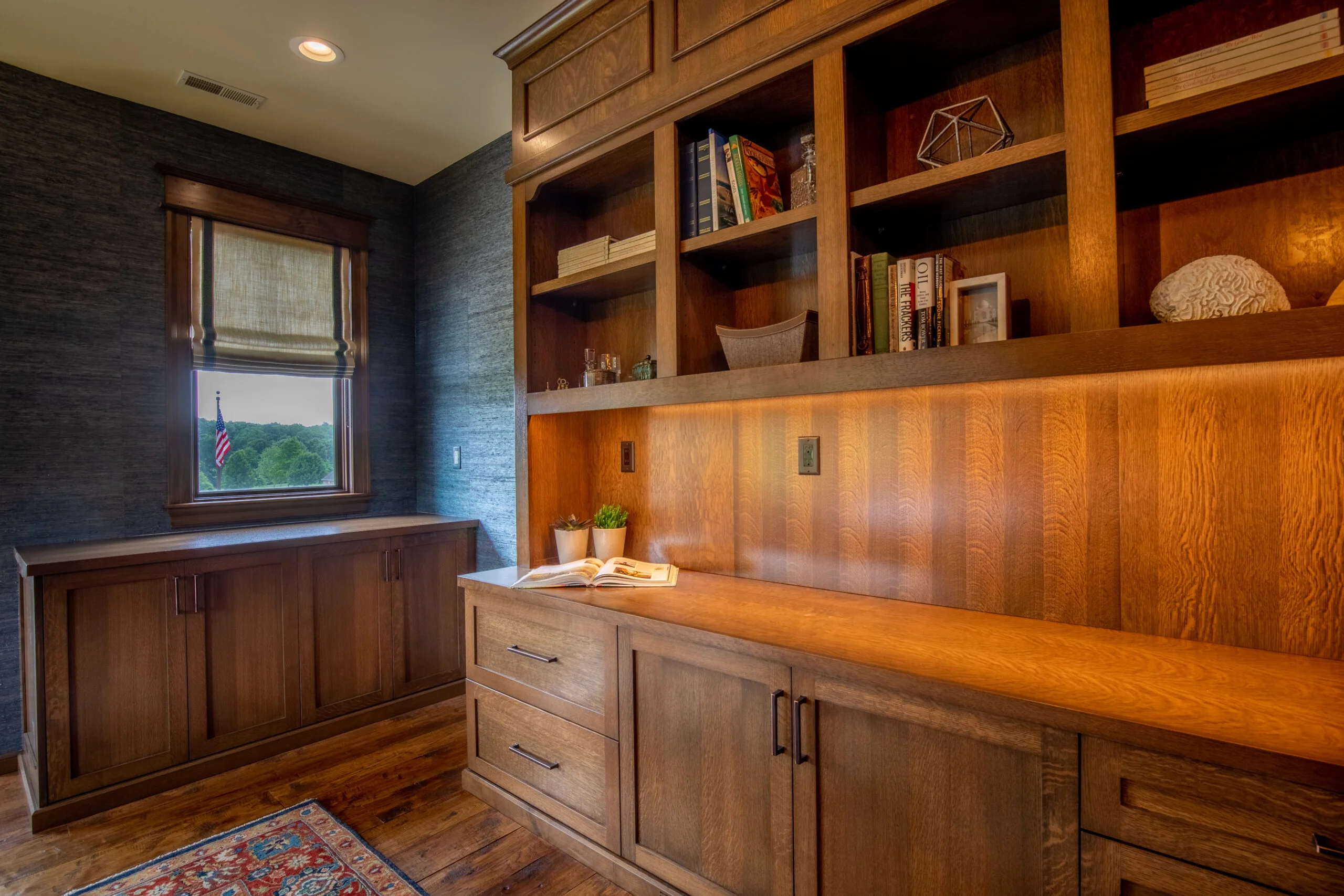 Built-in oak cabinetry with shelving, desk, and under-cabinet lighting. Window reveals US flag & green trees.
