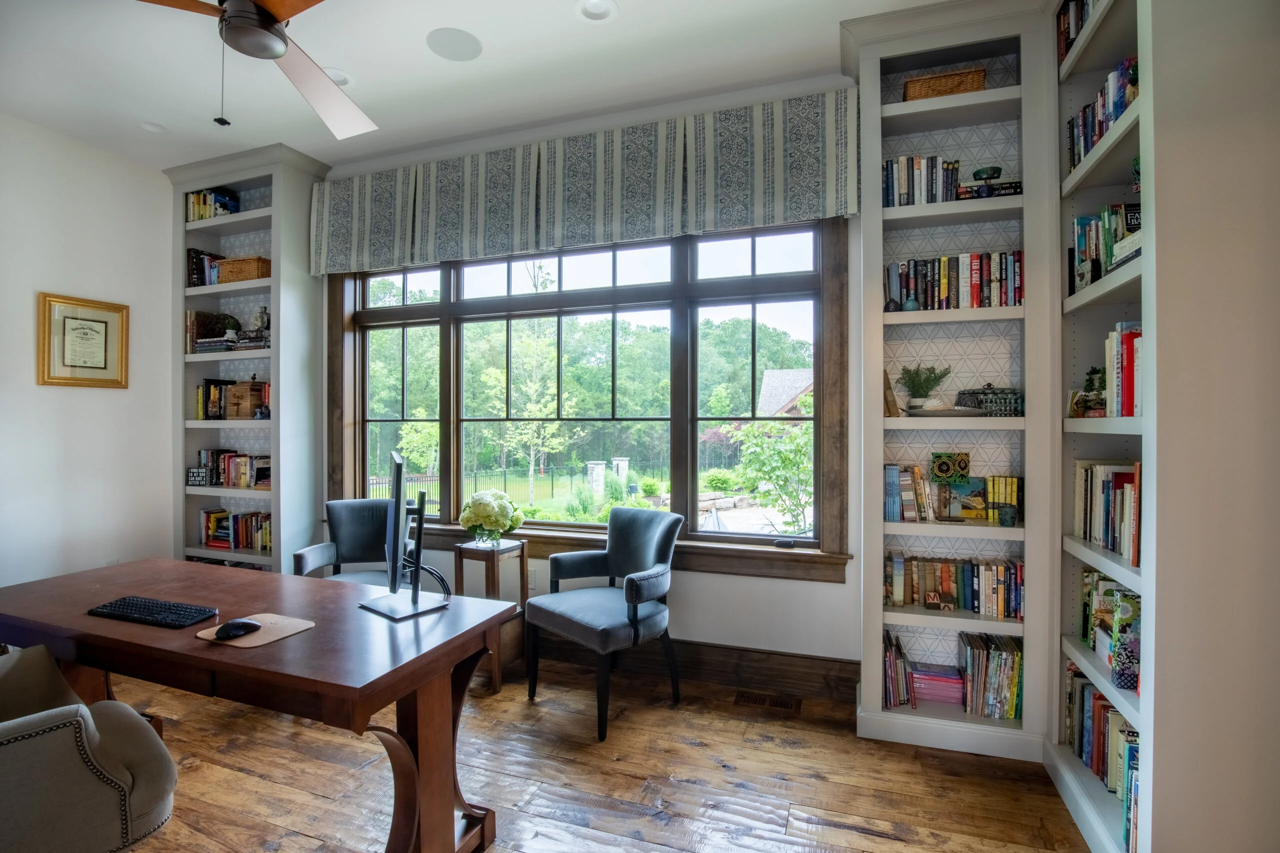 Home office with large wooden desk, grey upholstered chairs, built-in bookshelves, and large window overlooking trees.