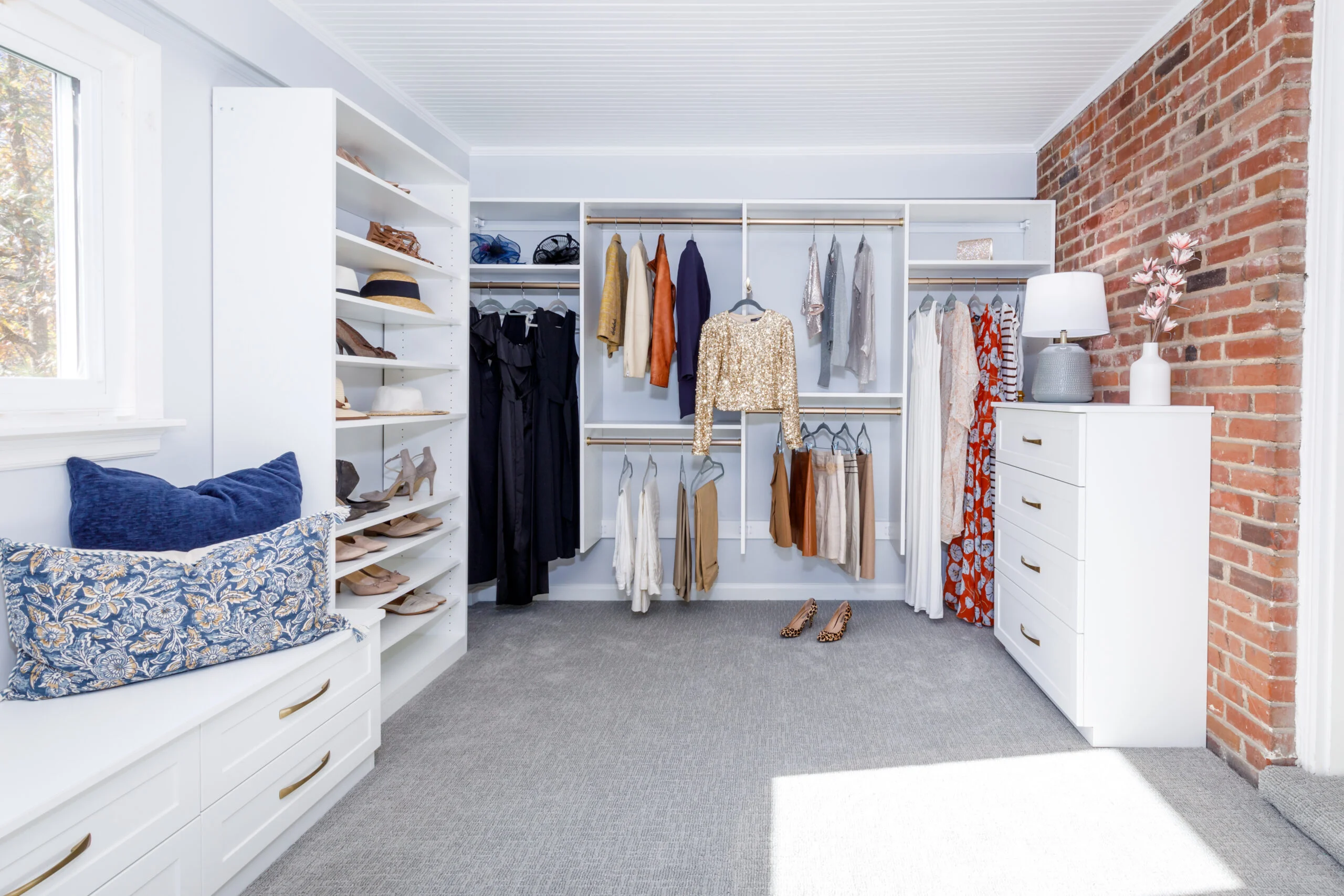 Bright, organized walk-in closet with white shelving, a brick accent wall, and a window overlooking autumn trees.