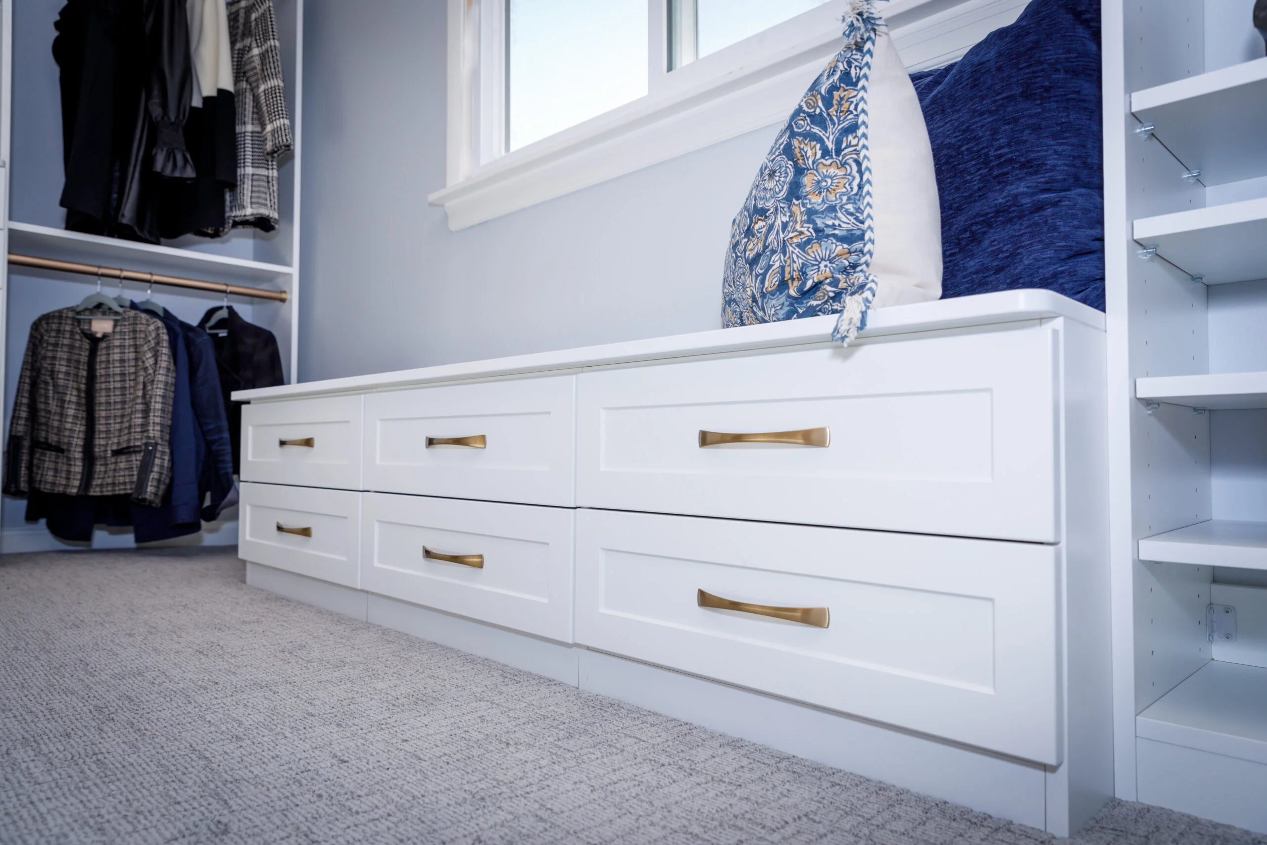 White built-in storage bench with gold handles, blue cushions, next to a window and hanging clothing in a closet.