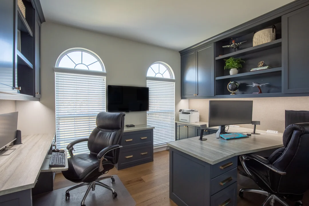 A stylish home office with dark blue cabinets, light wood counters, two black office chairs, arched windows, and decorative shelves.
