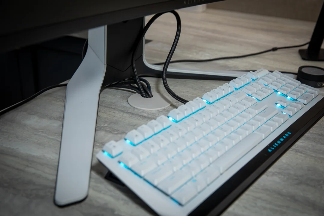 Alienware gaming keyboard with white keys and glowing cyan backlighting on a light wood desk.
