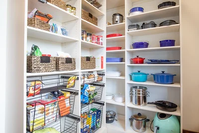 Organized pantry with white shelves, wicker baskets, wire racks, colorful cookware, and kitchen appliances.